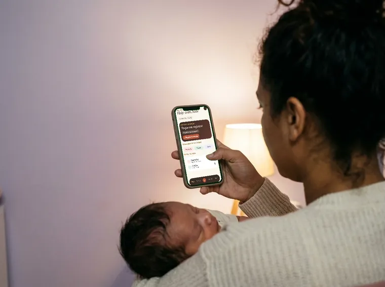 Mão segurando o celular com o polegar, enquanto o outro braço embala o bebê.