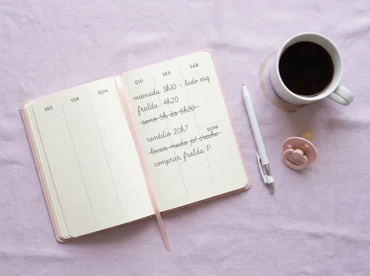 Caderno aberto com anotações riscadas, uma caneta, uma chupeta e um café frio.